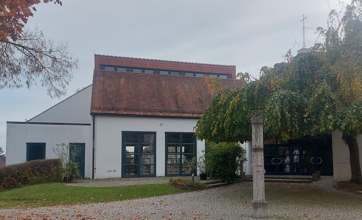 Pfarrzentrum mit Eingangstür und großer Fensterfront. Rechts neben der Tür steht ein großer Baum