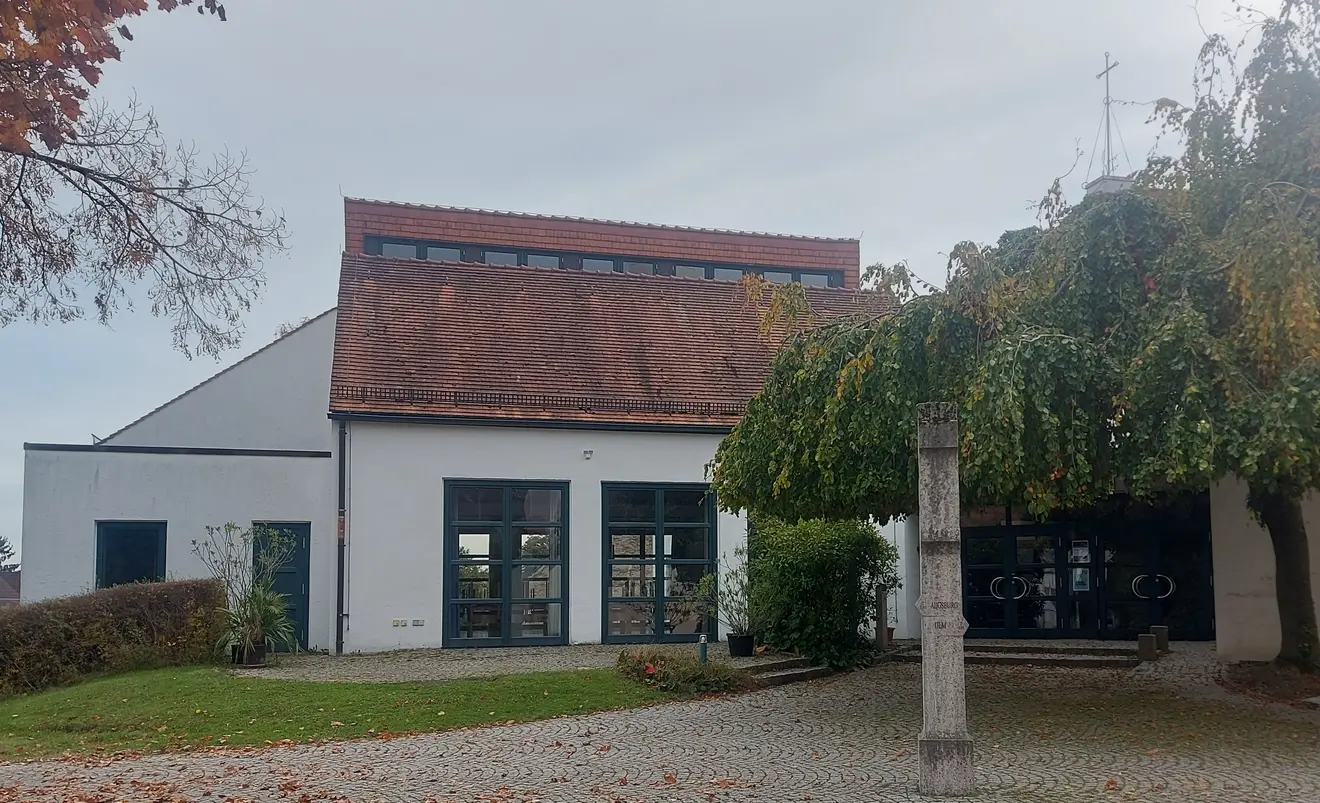 Pfarrzentrum mit Eingangstür und großer Fensterfront. Rechts neben der Tür steht ein großer Baum