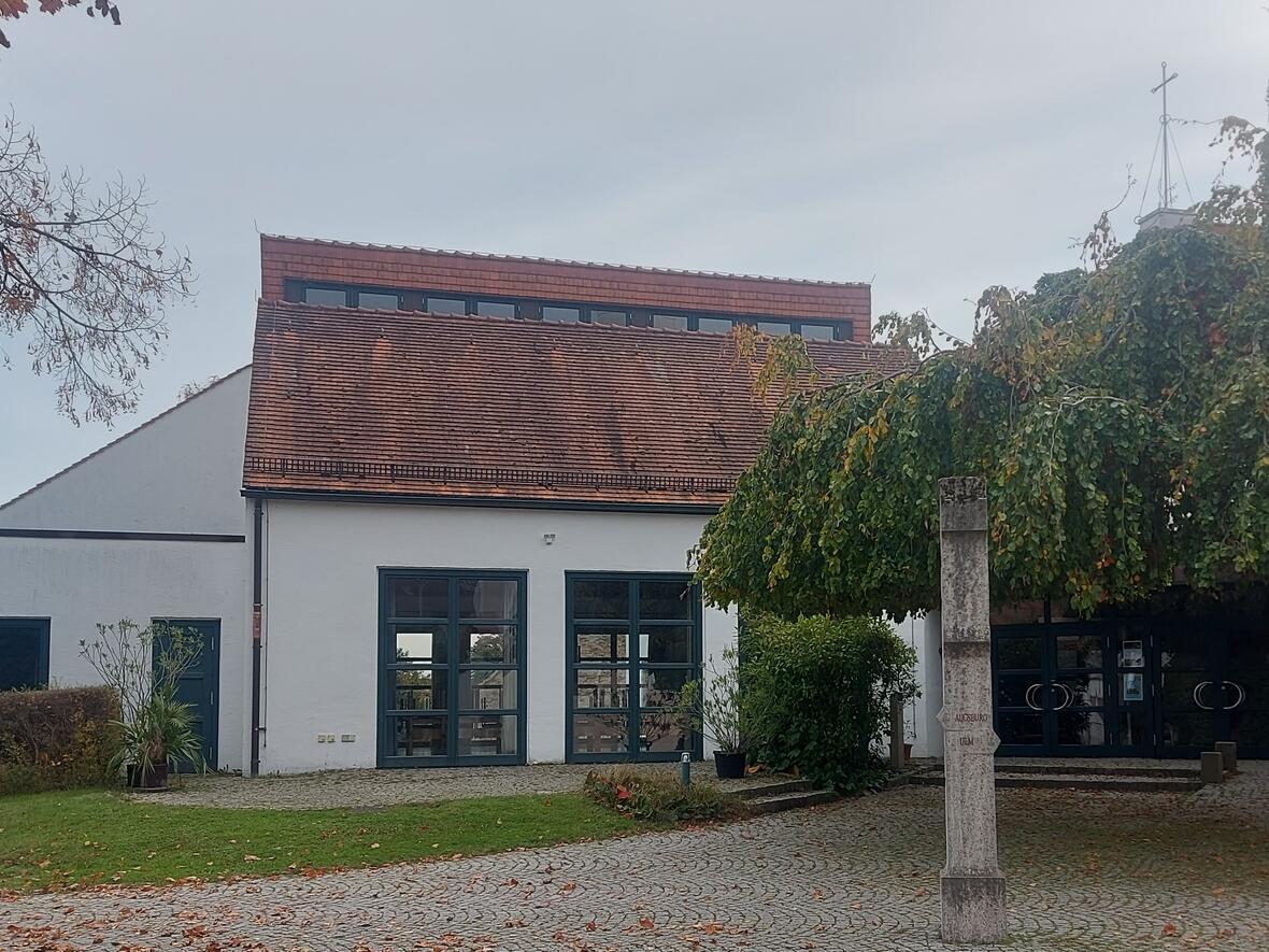 Pfarrzentrum mit Eingangstür und großer Fensterfront. Rechts neben der Tür steht ein großer Baum