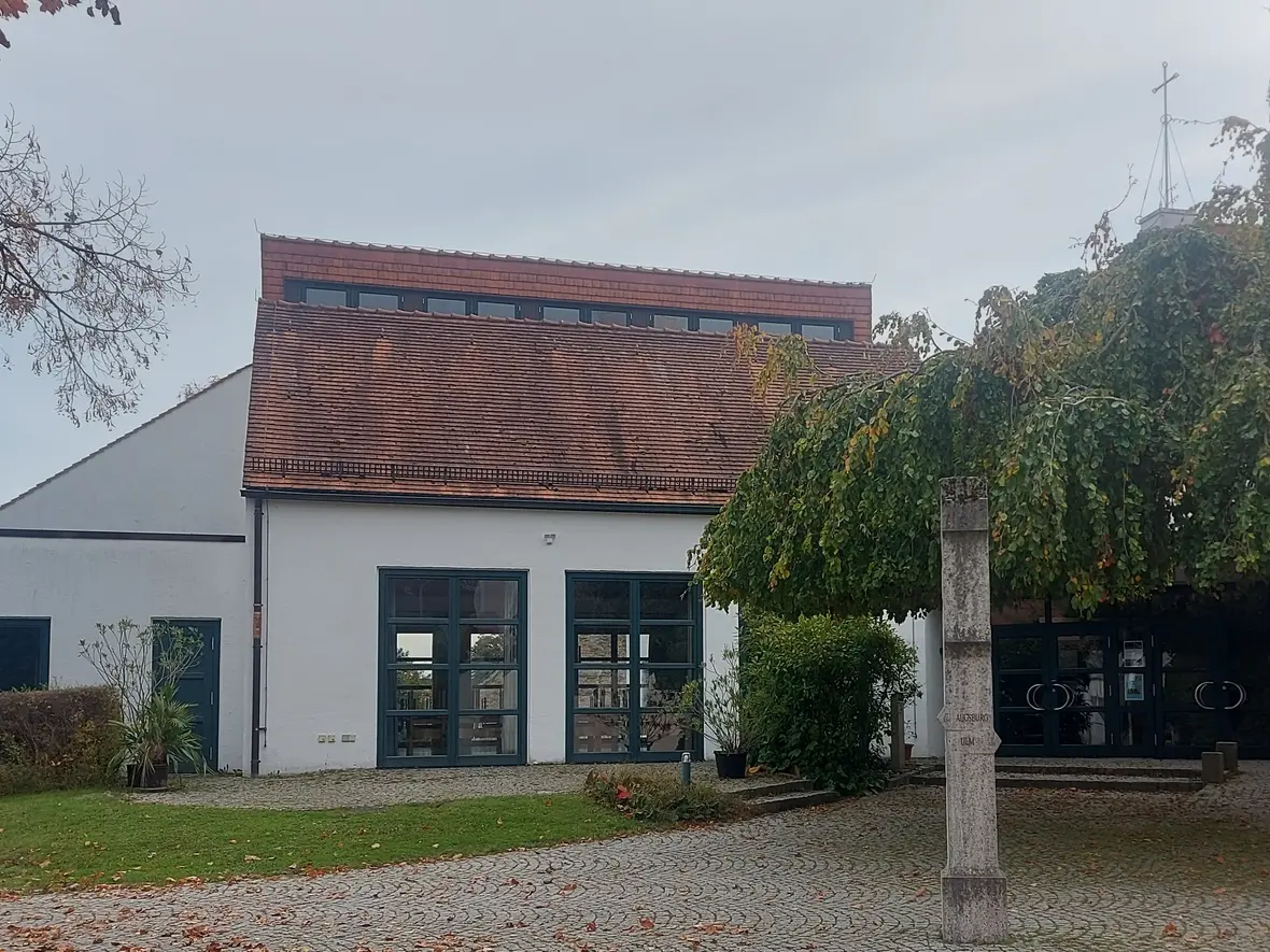 Pfarrzentrum mit Eingangstür und großer Fensterfront. Rechts neben der Tür steht ein großer Baum