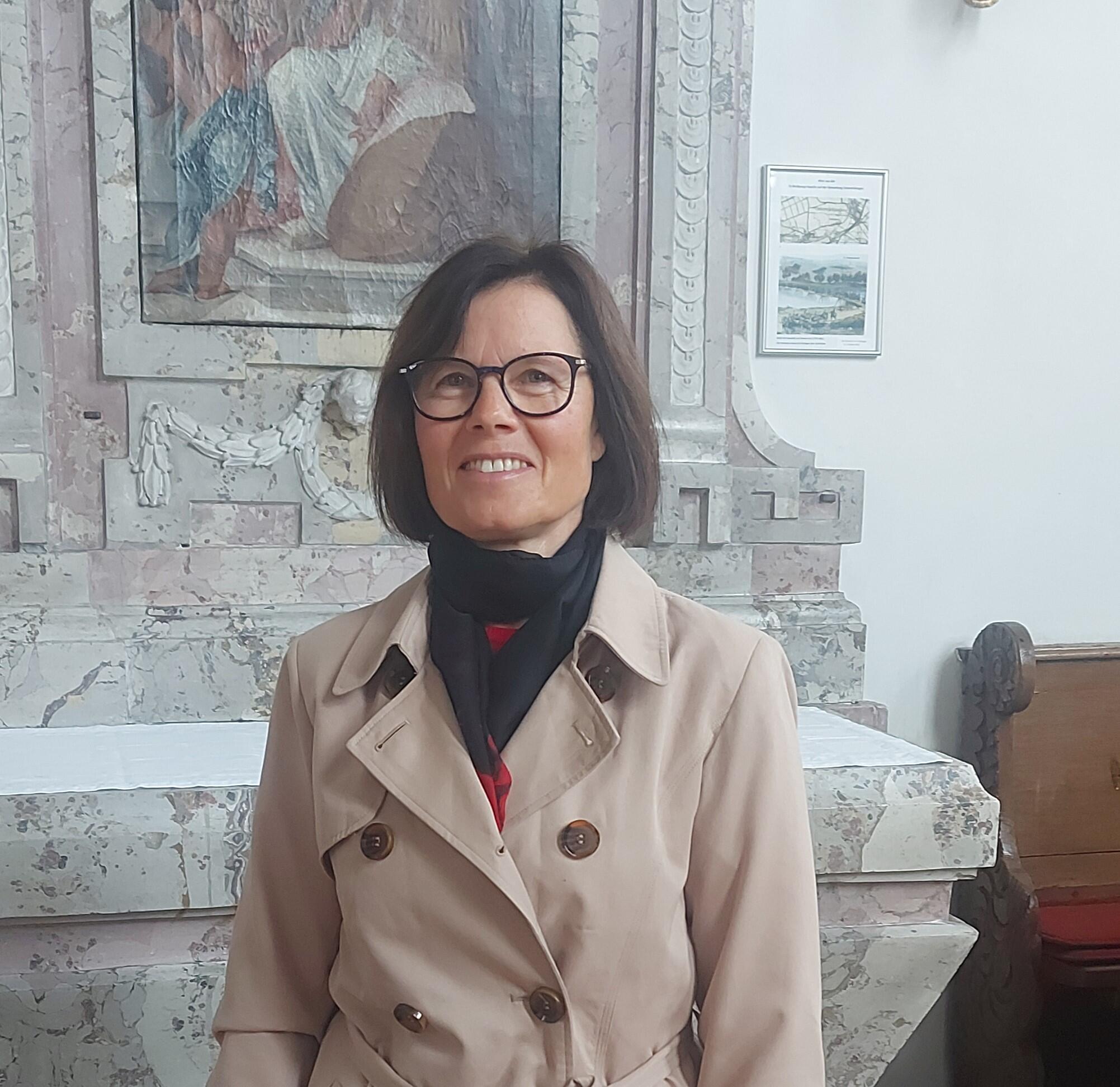 Frau lächelnd, lange dunkle Haare und Brille steht in der Kirche vor einem Seitenaltar
