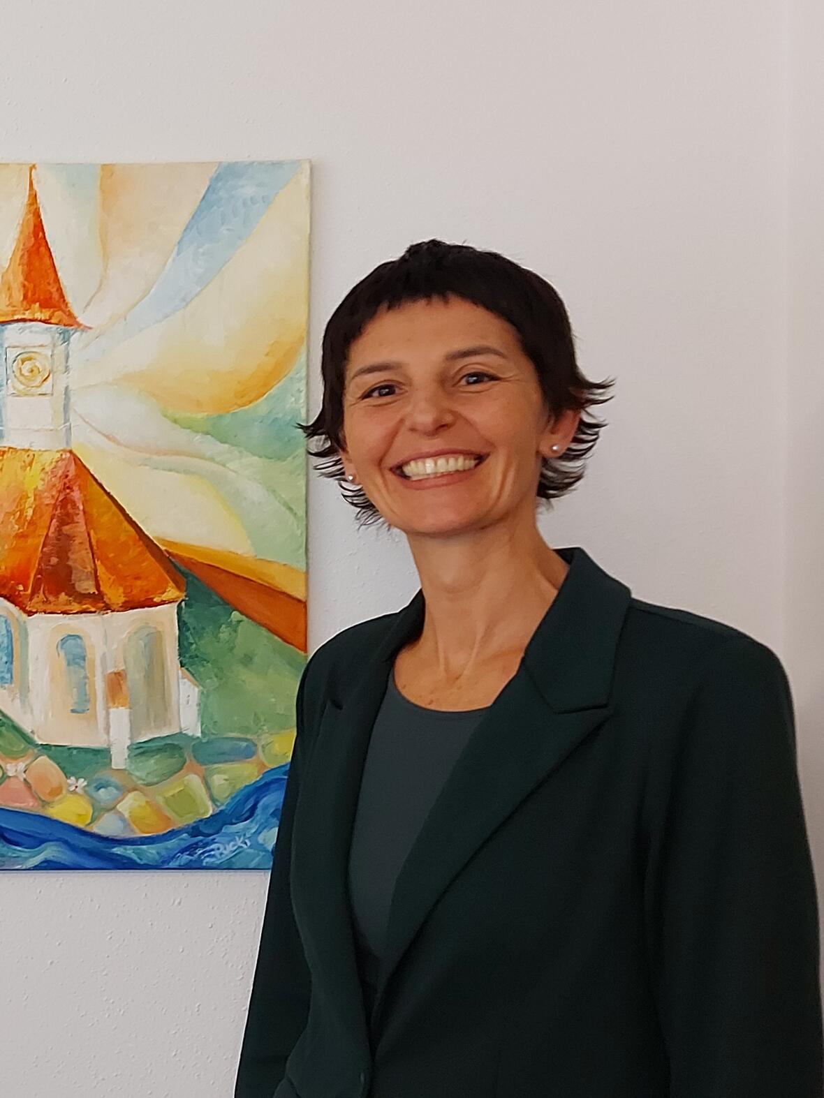 Jenny Moos Buchhaltung Frau steht lächelnd vor einem Bild von der Kirche St. Laurentius. Sie hat kurze schwarze Haare