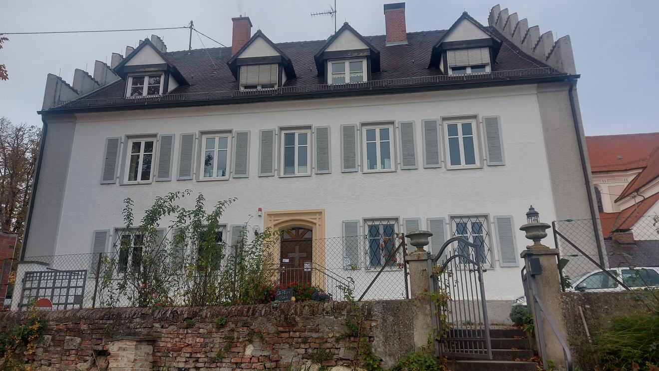 Pfarrhaus Oberelchingen mit hellem Fassadenanstrich und grünen Fensterläden. Garten mit Bepflanzung.