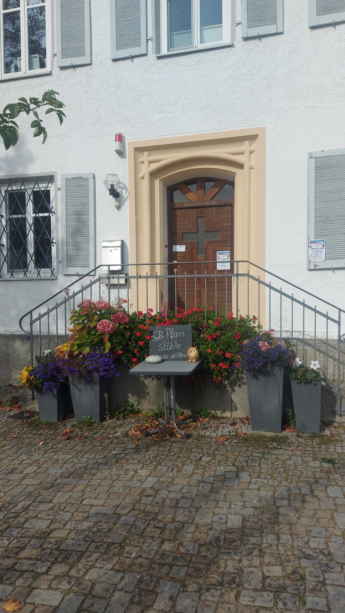 Pfarrhaus Oberelchingen mit blühenden Pflanzen vor der Haustür und einem Schild zur Pfarrstube.