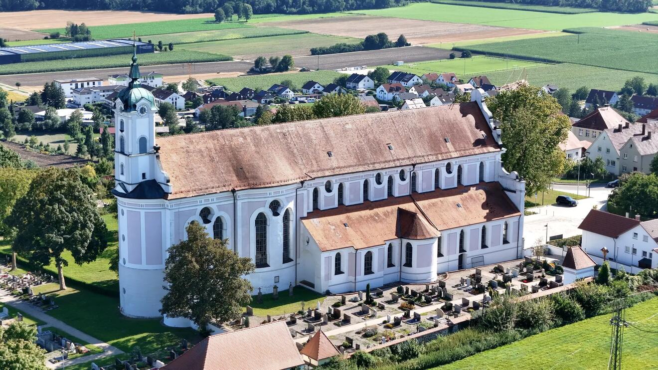 Kirche mit Grabsteinanlage im Vordergrund, umgeben von Feldern und Wohnhäusern.