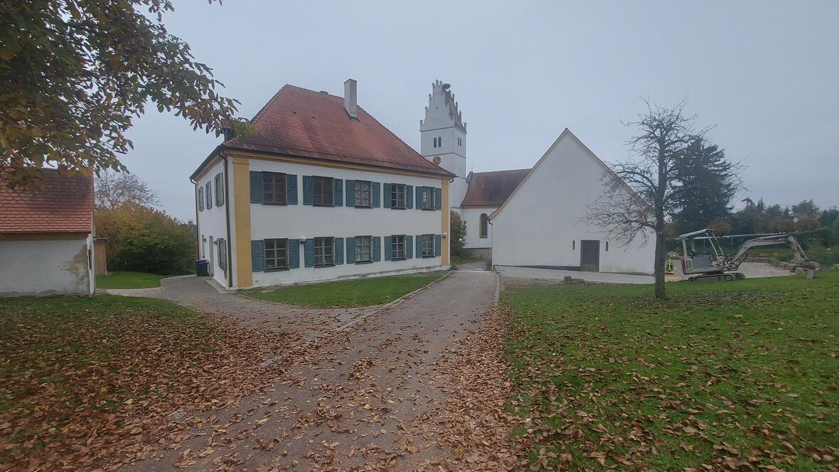 Historisches Gebäude mit roten Dächern, umgeben von Laubbäumen und einem kleinen Bagger im Vordergrund.