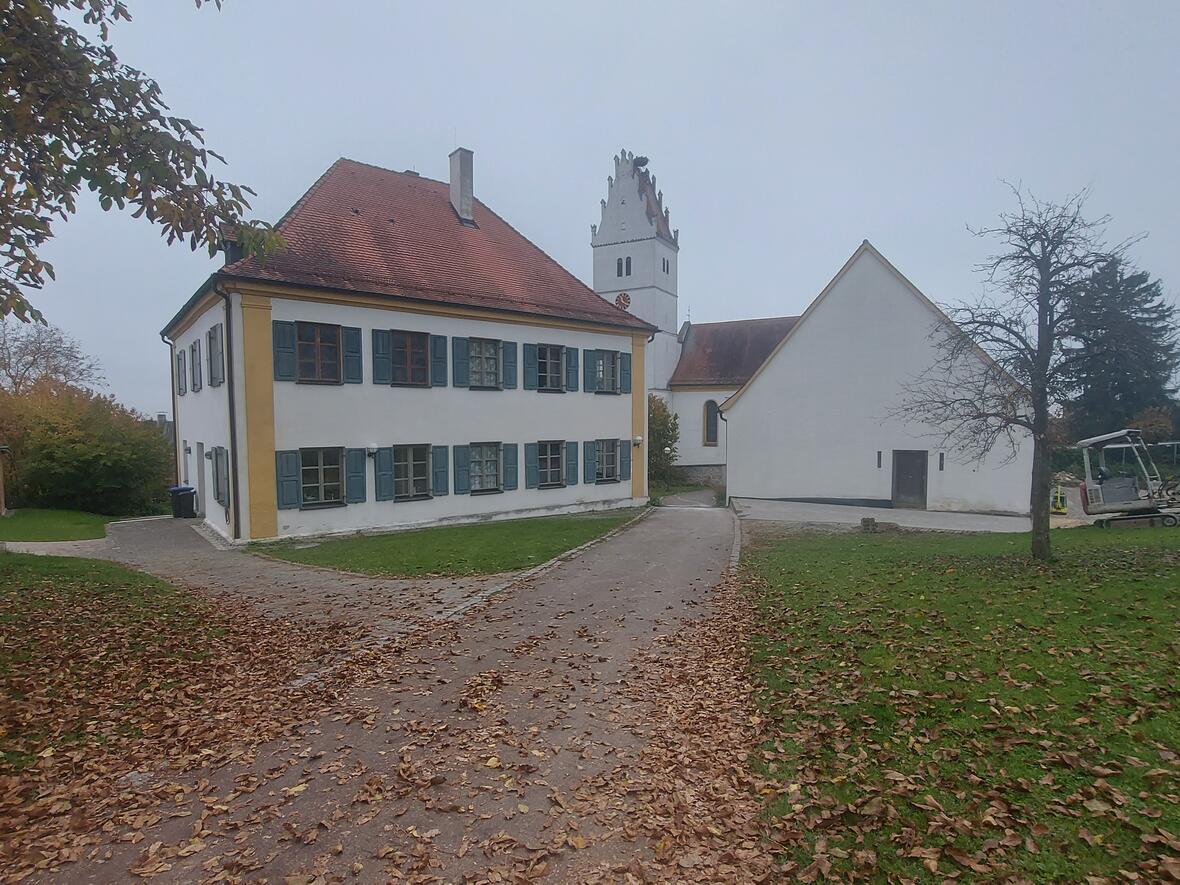 Historisches Gebäude mit roten Dächern, umgeben von Laubbäumen und einem kleinen Bagger im Vordergrund.