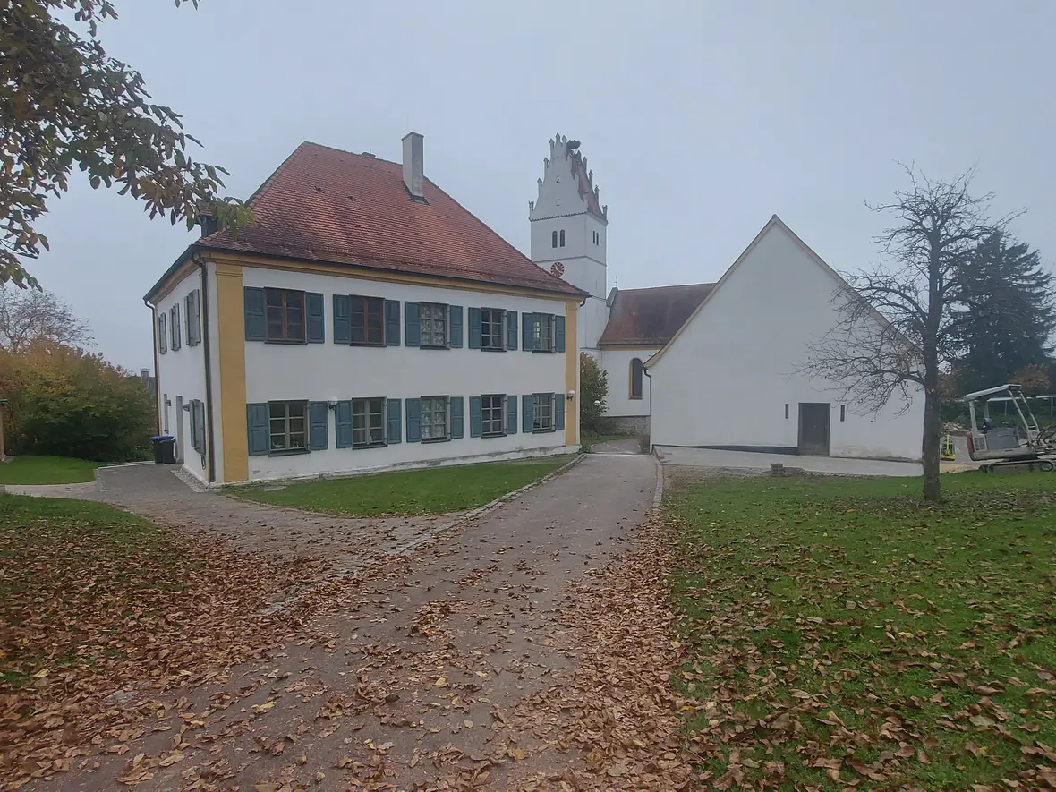 Historisches Gebäude mit roten Dächern, umgeben von Laubbäumen und einem kleinen Bagger im Vordergrund.