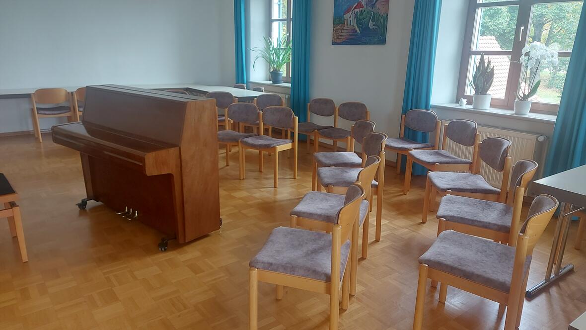 Ein Raum mit einem Klavier und mehreren Stühlen, umgeben von Pflanzen und Fenstern.