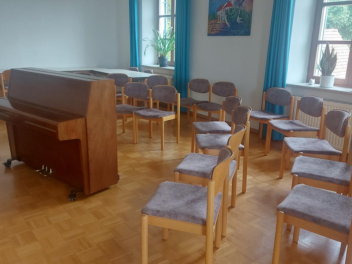 Ein Raum mit einem Klavier und mehreren Stühlen, umgeben von Pflanzen und Fenstern.