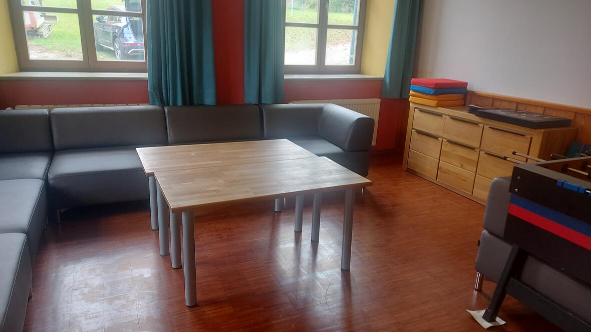 Gemütlicher Aufenthaltsraum mit grauem Sofa, Holztisch und bunten Matten. Fensterausblick ins Grüne.