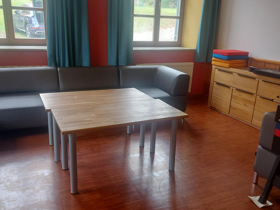 Gemütlicher Aufenthaltsraum mit grauem Sofa, Holztisch und bunten Matten. Fensterausblick ins Grüne.