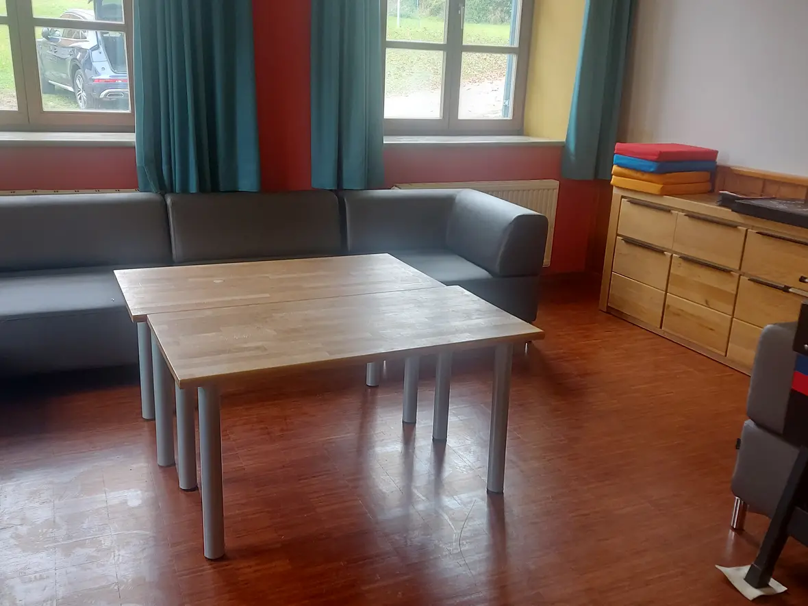 Gemütlicher Aufenthaltsraum mit grauem Sofa, Holztisch und bunten Matten. Fensterausblick ins Grüne.