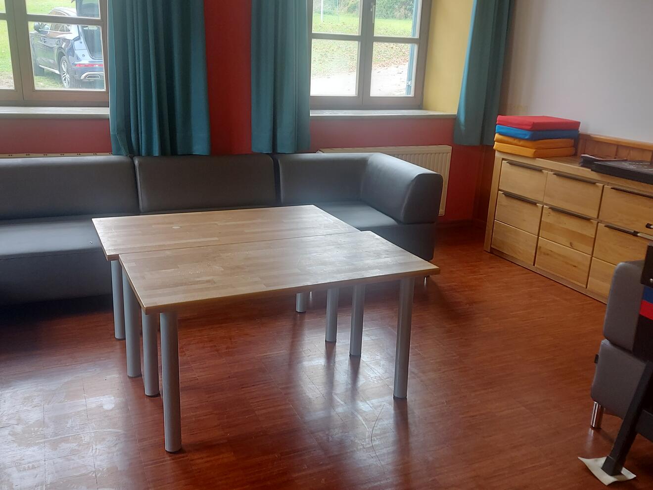 Gemütlicher Aufenthaltsraum mit grauem Sofa, Holztisch und bunten Matten. Fensterausblick ins Grüne.