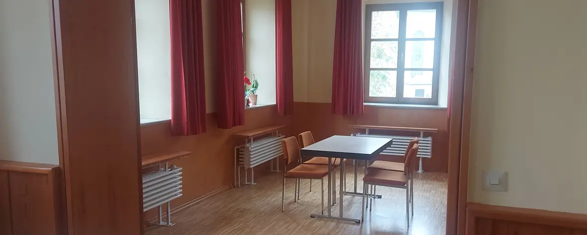 Gemütlicher Raum mit Tisch und Stühlen, großen Fenstern und roten Vorhängen.