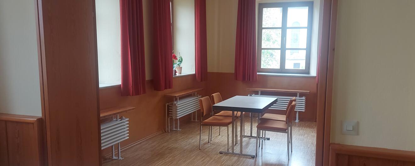Gemütlicher Raum mit Tisch und Stühlen, großen Fenstern und roten Vorhängen.