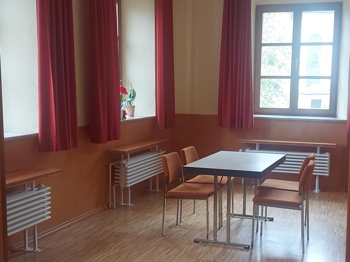 Gemütlicher Raum mit Tisch und Stühlen, großen Fenstern und roten Vorhängen.