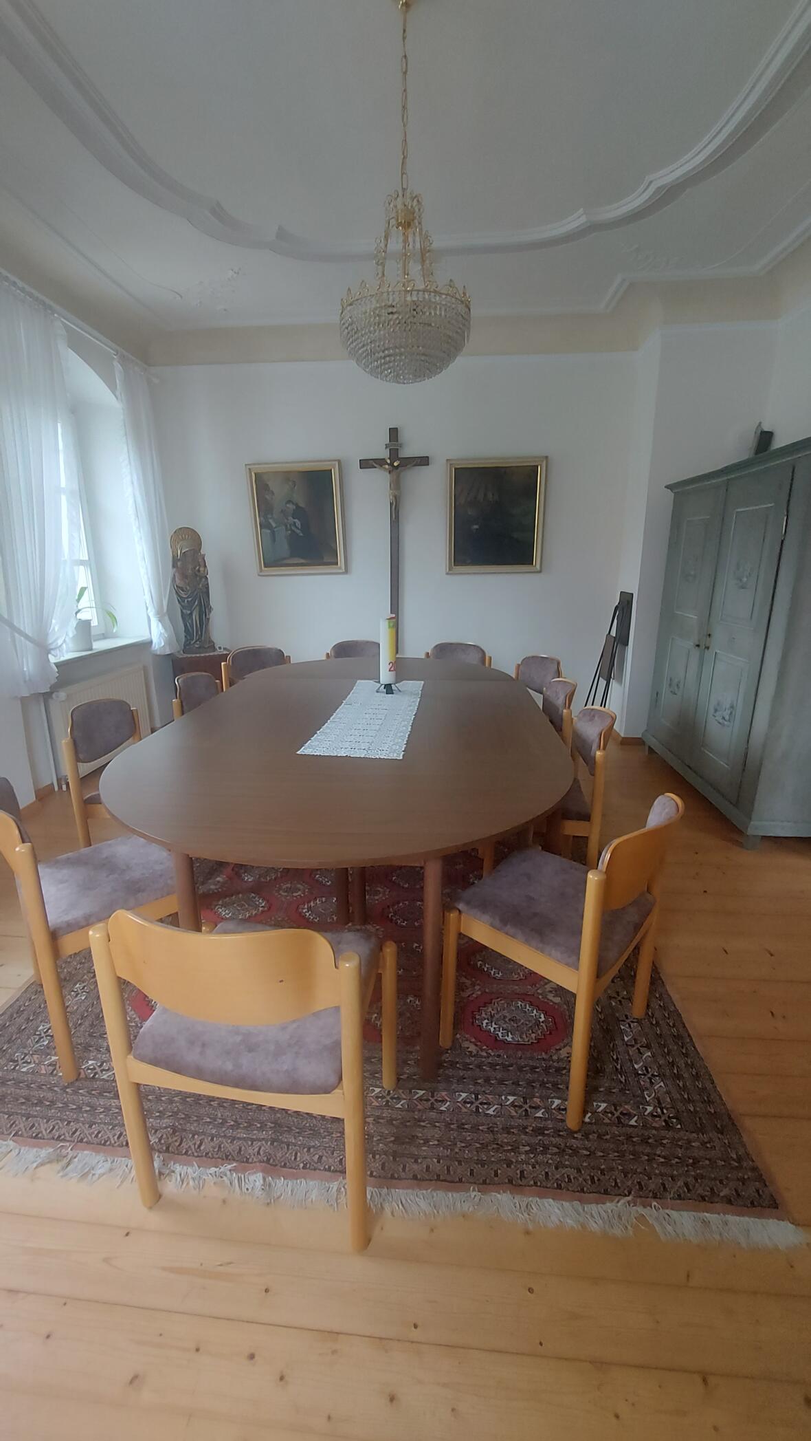 Ein ovaler Tisch mit Stühlen in einem Raum mit einem Kruzifix und zwei Bildern an der Wand.