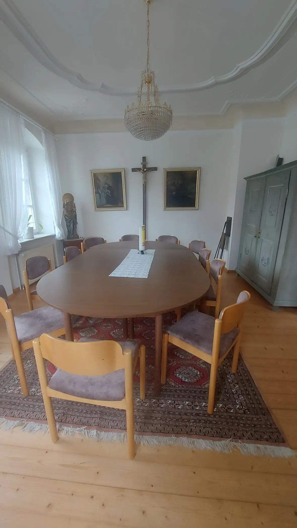 Ein ovaler Tisch mit Stühlen in einem Raum mit einem Kruzifix und zwei Bildern an der Wand.