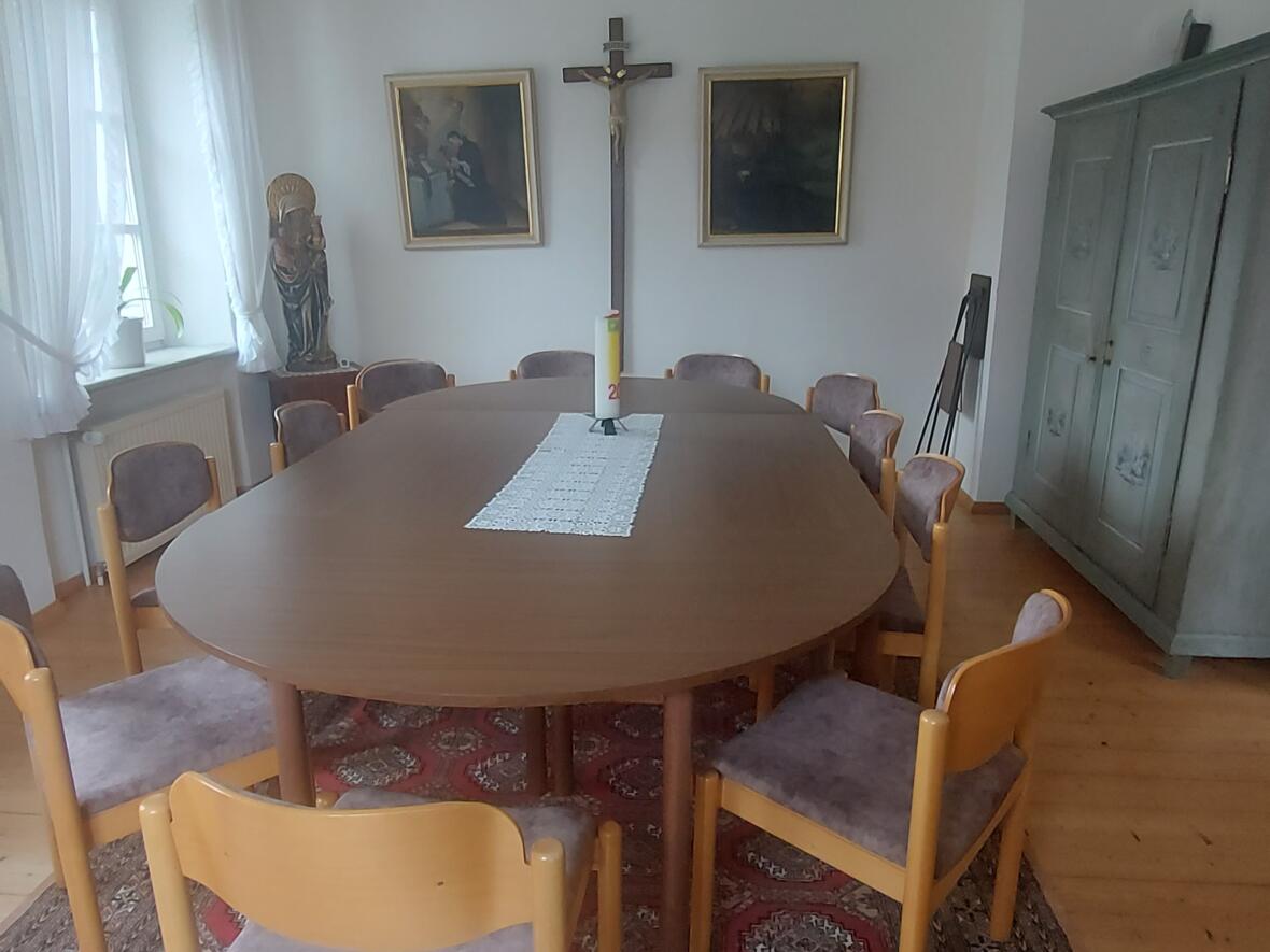 Ein ovaler Tisch mit Stühlen in einem Raum mit einem Kruzifix und zwei Bildern an der Wand.