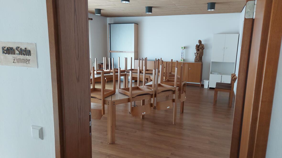 Ein heller Raum. In der Mitte stehen mehrere Tische. Auf jedem Tisch liegen Stühle mit den Beinen nach oben. Rechts im Raum stehen Schränke und eine Holz-Figur. An der Wand außen am Türrahmen steht: „Edith Stein Zimmer“.