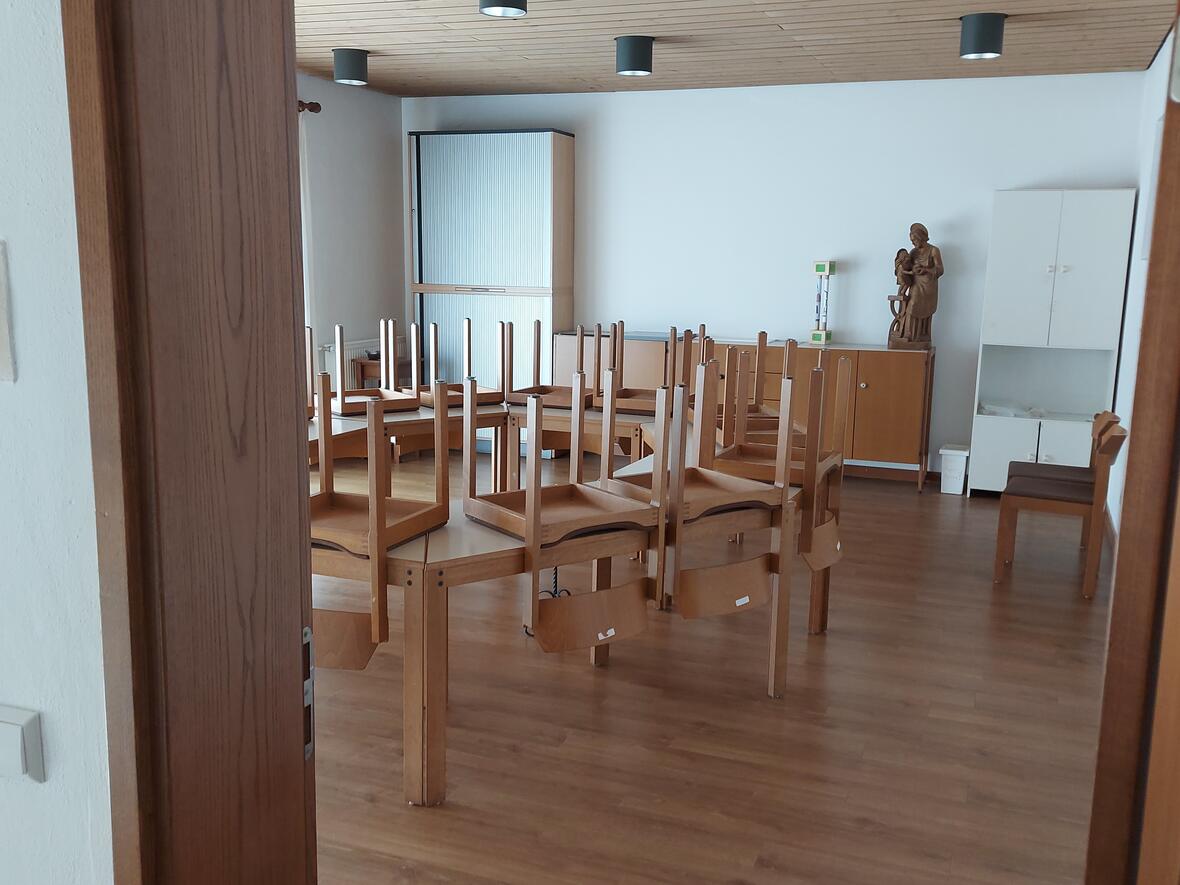 Edith-Stein-Zimmer Ein heller Raum. In der Mitte stehen mehrere Tische. Auf jedem Tisch liegen Stühle mit den Beinen nach oben. Rechts im Raum stehen Schränke und eine Holz-Figur. An der Wand außen am Türrahmen steht: „Edith Stein Zimmer“.