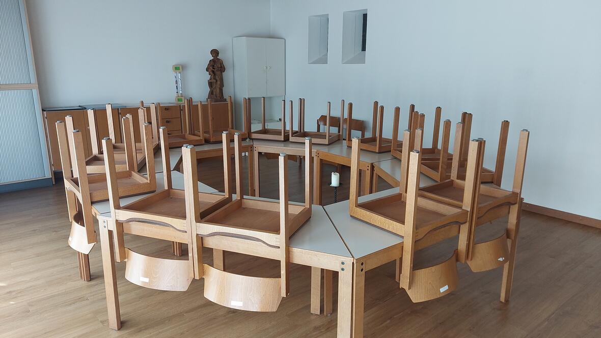Edith-Stein-Zimmer Viele Holztische stehen im Kreis. Auf jedem Tisch liegen Stühle mit den Beinen nach oben. Im Hintergrund stehen Schränke und eine Holz-Figur. Auf dem Boden steht eine weiße Kerze in einem schwarzen Kerzenhalter.