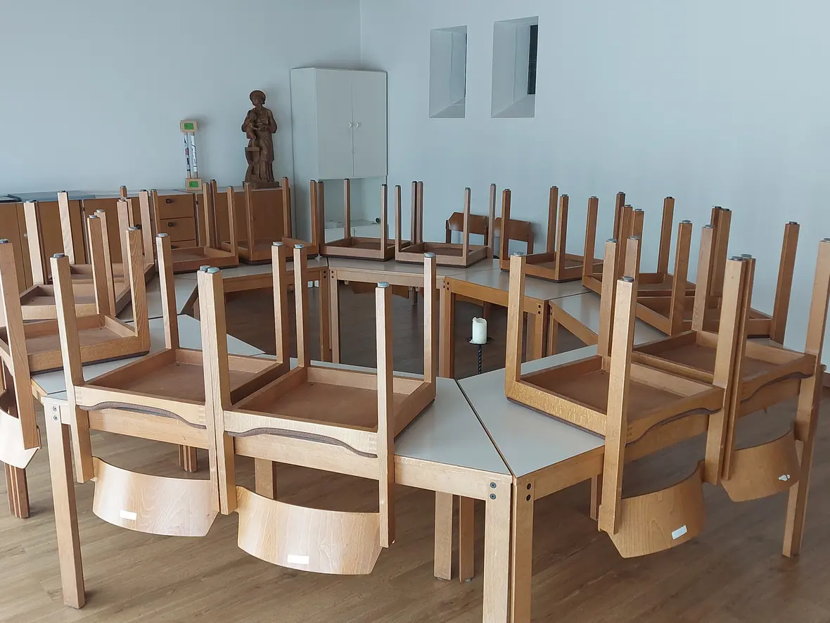 Viele Holztische stehen im Kreis. Auf jedem Tisch liegen Stühle mit den Beinen nach oben. Im Hintergrund stehen Schränke und eine Holz-Figur. Auf dem Boden steht eine weiße Kerze in einem schwarzen Kerzenhalter.