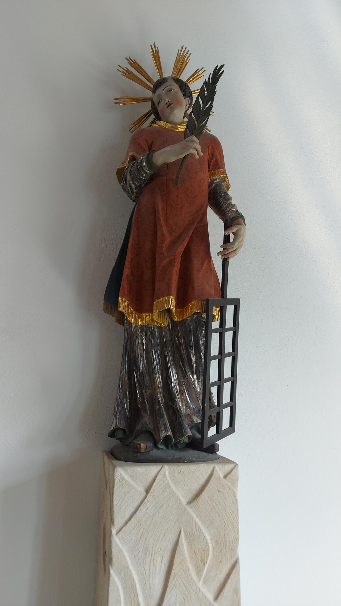 Eine bemalte Figur steht auf einem hellen Sockel. Die Figur trägt ein rotes Gewand mit goldenen Rändern. In der linken Hand hält sie ein Gitter. In der rechten Hand hält sie einen langen Zweig. Hinter dem Kopf sind goldene Strahlen zu sehen.