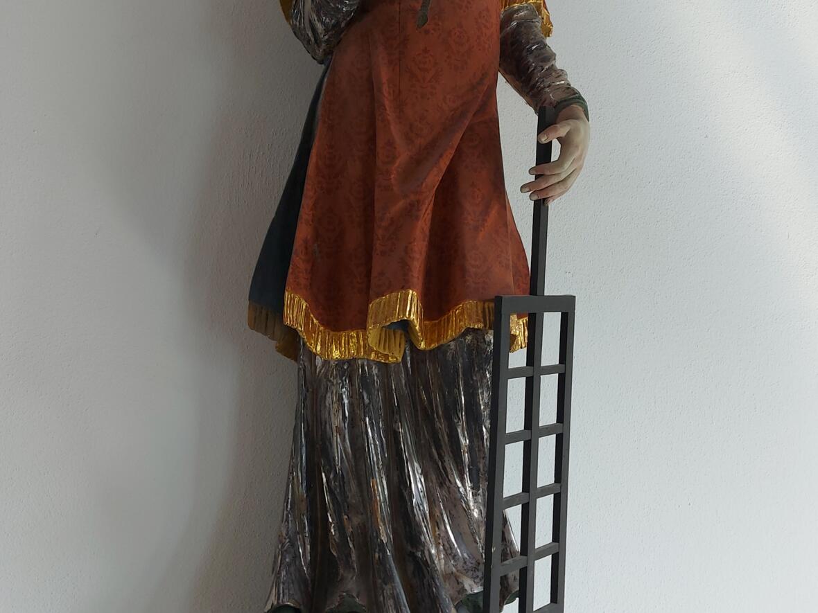 Heiliger Laurentius Eine bemalte Figur steht auf einem hellen Sockel. Die Figur trägt ein rotes Gewand mit goldenen Rändern. In der linken Hand hält sie ein Gitter. In der rechten Hand hält sie einen langen Zweig. Hinter dem Kopf sind goldene Strahlen zu sehen.