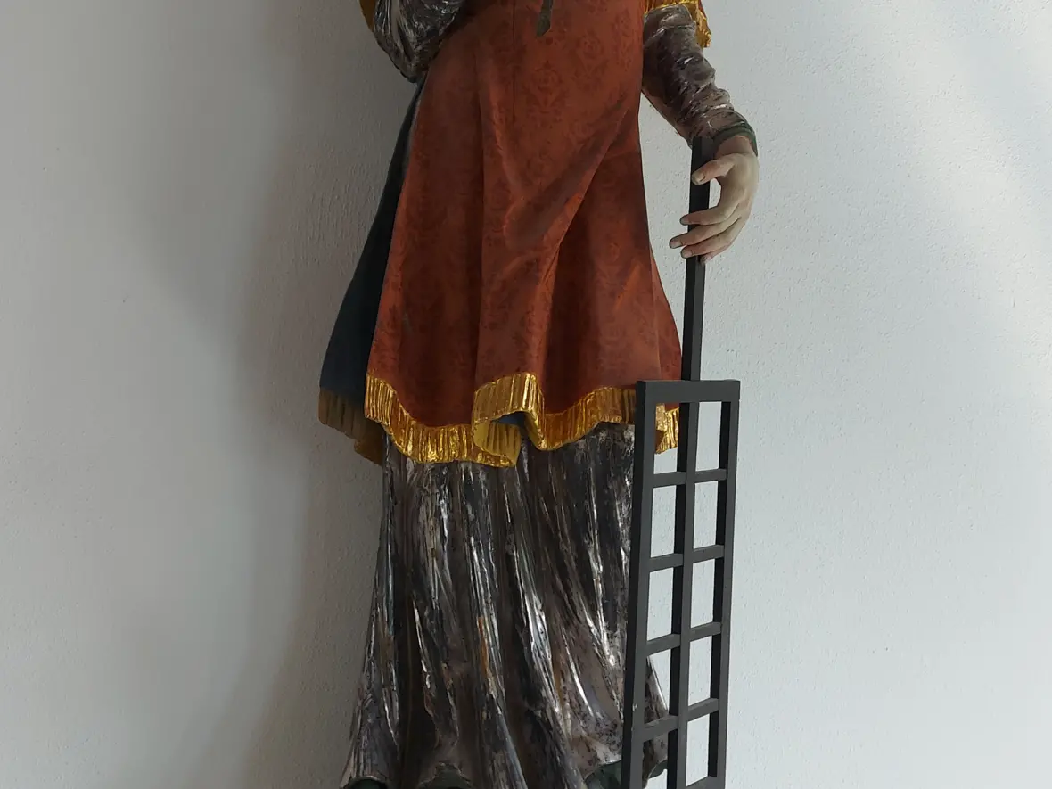 Eine bemalte Figur steht auf einem hellen Sockel. Die Figur trägt ein rotes Gewand mit goldenen Rändern. In der linken Hand hält sie ein Gitter. In der rechten Hand hält sie einen langen Zweig. Hinter dem Kopf sind goldene Strahlen zu sehen.