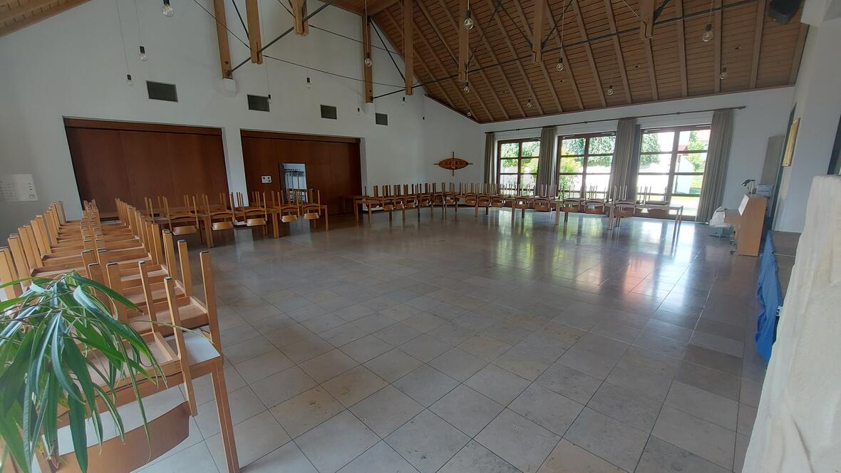 Ein großer Saal mit vielen Fenstern und hellem Boden. An den Wänden stehen viele Tische. Auf jedem Tisch liegen Stühle mit den Beinen nach oben. Links vorne steht eine grüne Pflanze. Rechts im Raum steht ein Klavier. Die Decke ist hoch und hat Holzbalken.