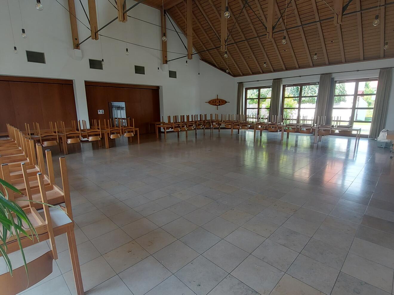 Ein großer Saal mit vielen Fenstern und hellem Boden. An den Wänden stehen viele Tische. Auf jedem Tisch liegen Stühle mit den Beinen nach oben. Links vorne steht eine grüne Pflanze. Rechts im Raum steht ein Klavier. Die Decke ist hoch und hat Holzbalken.