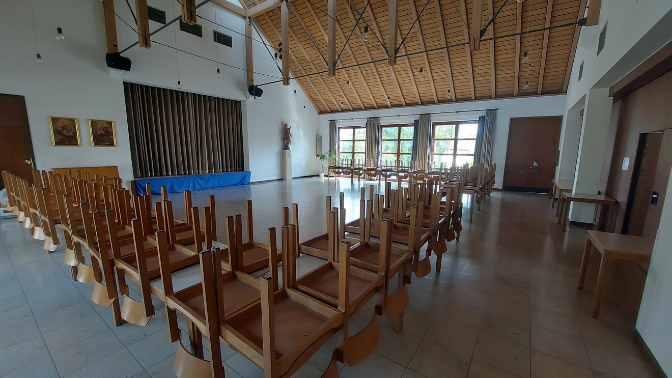 Ein großer Saal mit vielen Tischen. Auf den Tischen liegen Stühle mit den Beinen nach oben. Der Raum hat eine hohe Decke mit Holzbalken. Rechts und vorne sind große Fenster mit Vorhängen. An einer Wand hängen zwei Bilder. Daneben steht eine Figur auf einem Sockel. In der Mitte der Wand befindet sich ein großer Durchgang mit einem braunen Vorhang dahinter.
