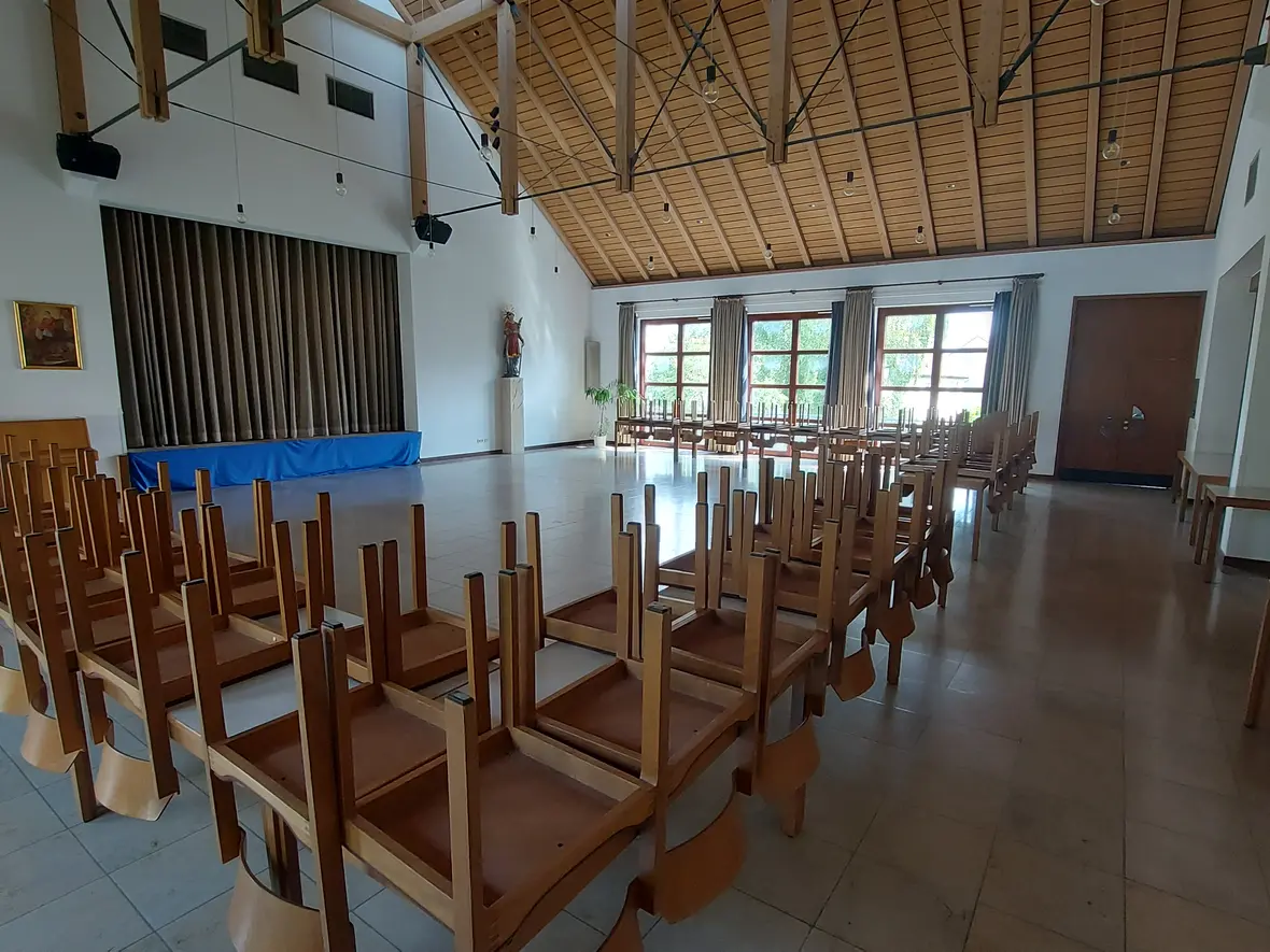 Ein großer Saal mit vielen Tischen. Auf den Tischen liegen Stühle mit den Beinen nach oben. Der Raum hat eine hohe Decke mit Holzbalken. Rechts und vorne sind große Fenster mit Vorhängen. An einer Wand hängen zwei Bilder. Daneben steht eine Figur auf einem Sockel. In der Mitte der Wand befindet sich ein großer Durchgang mit einem braunen Vorhang dahinter.