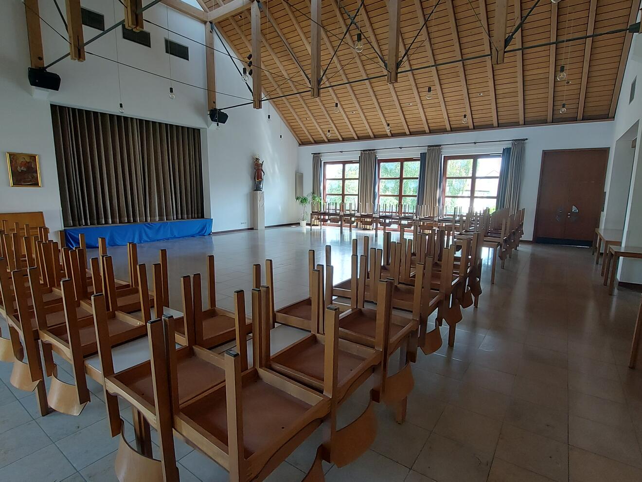 Ein großer Saal mit vielen Tischen. Auf den Tischen liegen Stühle mit den Beinen nach oben. Der Raum hat eine hohe Decke mit Holzbalken. Rechts und vorne sind große Fenster mit Vorhängen. An einer Wand hängen zwei Bilder. Daneben steht eine Figur auf einem Sockel. In der Mitte der Wand befindet sich ein großer Durchgang mit einem braunen Vorhang dahinter.