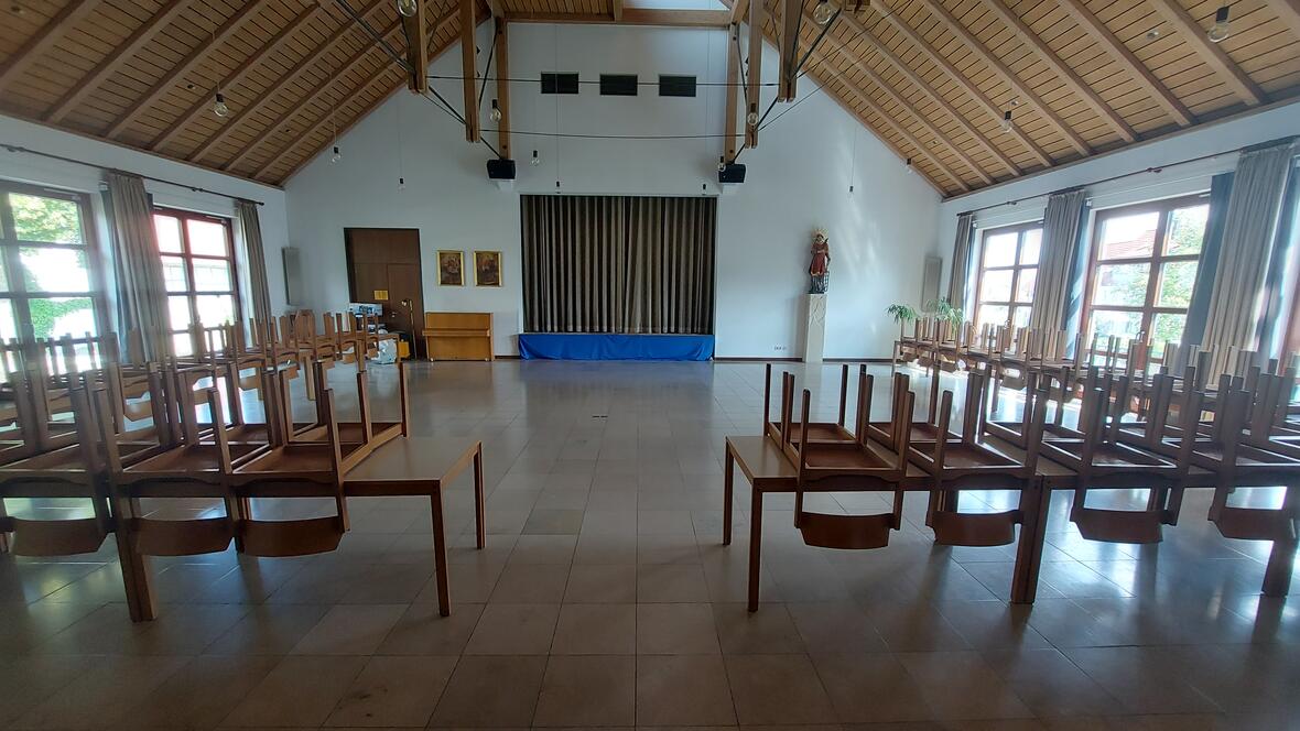 Ein großer Saal mit vielen Tischen. Auf den Tischen liegen Stühle mit den Beinen nach oben. Der Saal hat eine hohe Decke mit Holz-Balken. Vorne an der Wand ist ein großer Durchgang mit braunem Vorhang. Daneben stehen eine Figur auf einem Sockel und ein Klavier. Links hängen zwei Bilder an der Wand. Rechts und links sind große Fenster mit braunen Vorhängen.