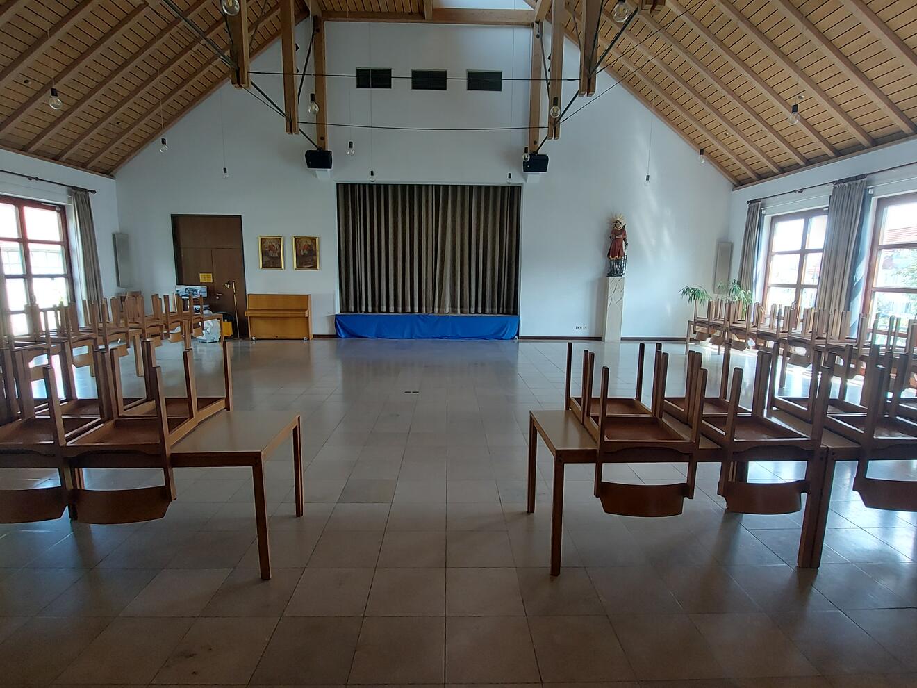 Ein großer Saal mit vielen Tischen. Auf den Tischen liegen Stühle mit den Beinen nach oben. Der Saal hat eine hohe Decke mit Holz-Balken. Vorne an der Wand ist ein großer Durchgang mit braunem Vorhang. Daneben stehen eine Figur auf einem Sockel und ein Klavier. Links hängen zwei Bilder an der Wand. Rechts und links sind große Fenster mit braunen Vorhängen.
